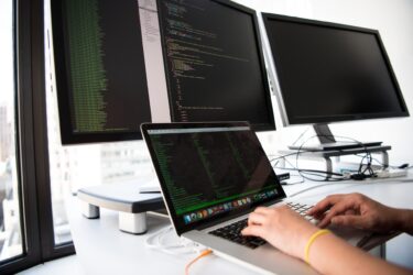 A person working on a laptop in front of two monitors, utilizing 2.0 and ASP.NET technologies.