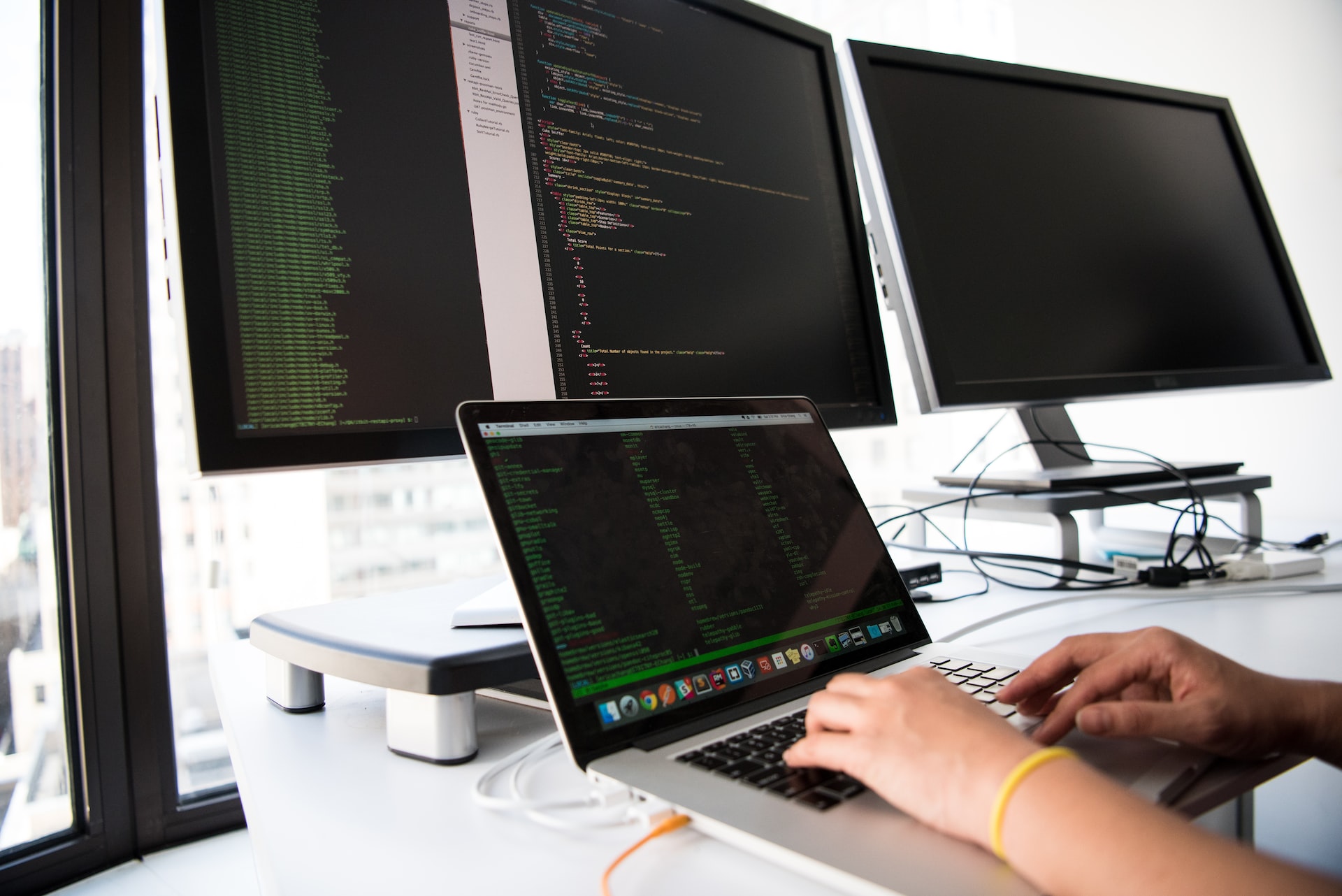 A person working on a laptop in front of two monitors, utilizing 2.0 and ASP.NET technologies.