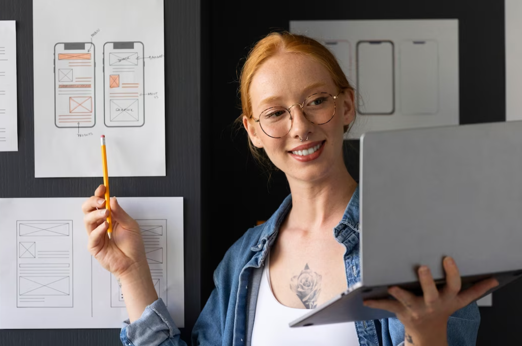 woman in glasses holding the laptop in one hand and pointing at the sheet pinned on the wall